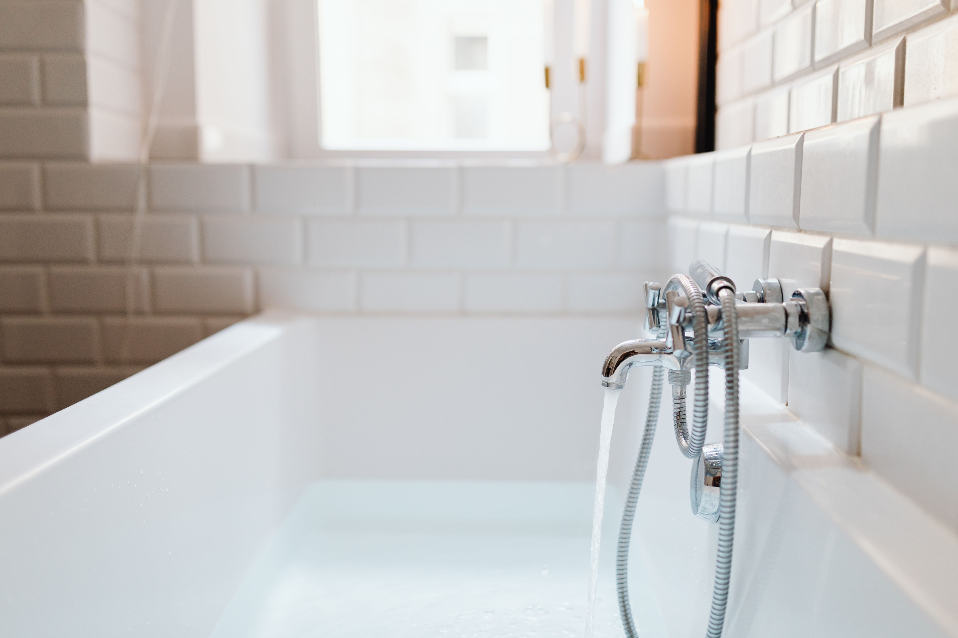 Bright white-tiled bathroom with a freshly filled tub and running chrome faucet after sanitizing service